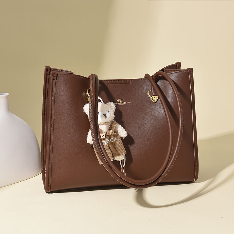 A professional product photo of a dark brown faux leather tote bag with a small white bear bag charm, resting on a light beige background next to a white vase